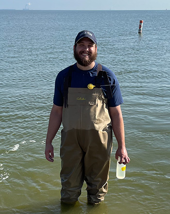 A man standing in a shallow part of Lake Erie, holding a bottle and smiling at the camera.